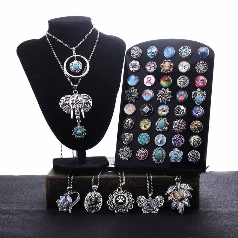 May include: A set of silver charm necklaces with various pendants, including an elephant, owl, heart, paw print, and leaf. The necklaces are displayed on a black stand with a variety of colorful snap buttons.
