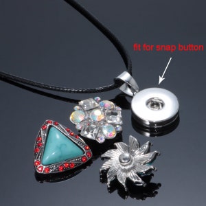 May include: A silver necklace with a black cord and a silver pendant that fits a snap button. The pendant is surrounded by three decorative snap buttons, one with turquoise stones, one with clear rhinestones, and one with a silver flower design.