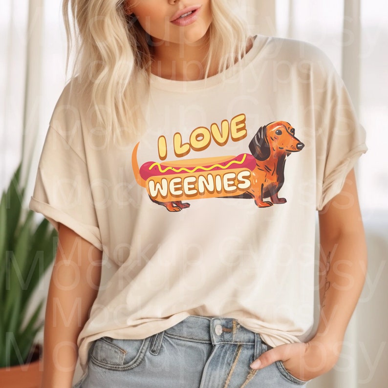 May include: A beige t-shirt featuring a dachshund dog dressed as a hot dog. The text on the shirt reads "I love weenies".