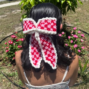 May include: A crochet hair bow featuring a checkered pattern in shades of red and light pink, finished with a white crocheted border. The bow has two long, dangling tails and is constructed with a textured yarn stitch throughout the design.