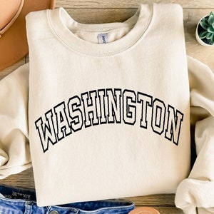 May include: A cream-colored sweatshirt with the word "WASHINGTON" printed in black across the front. The sweatshirt is laid out on a wooden surface with a brown hat and a small potted plant.