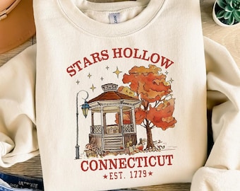May include: Cream-colored sweatshirt with a graphic of a gazebo, autumn tree, and the text "STARS HOLLOW CONNECTICUT EST. 1779". The design is in shades of brown, orange, and red. The sweatshirt is paired with blue jeans, a tan hat, and brown sandals.