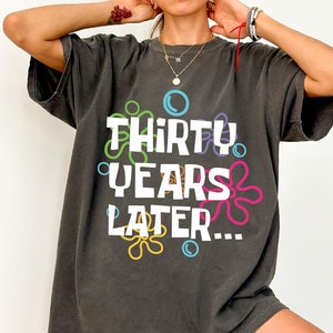 Puede incluir: Una camiseta de gran tama&ntilde;o de color gris oscuro con el texto "Thirty Years Later..." en blanco, rodeada de flores y burbujas de dibujos animados de colores.