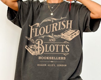 May include: A charcoal gray short-sleeve t-shirt featuring a beige graphic print that reads Flourish and Blotts Booksellers Diagon Alley, London, accompanied by illustrations of two books and a quill pen in a decorative layout.