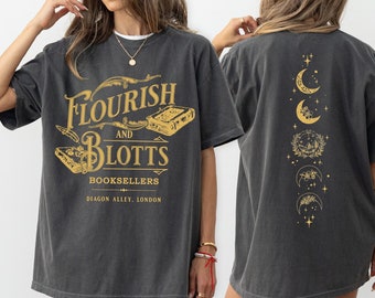 May include: A charcoal gray short-sleeve t-shirt featuring gold graphics. The front displays the text Flourish and Blotts Booksellers Diagon Alley, London with book illustrations. The back features a vertical design of five crescent moons.