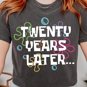 May include: A black cropped t-shirt with the text "TWENTY YEARS LATER..." in white lettering. The text is surrounded by colorful, cartoon-like shapes.