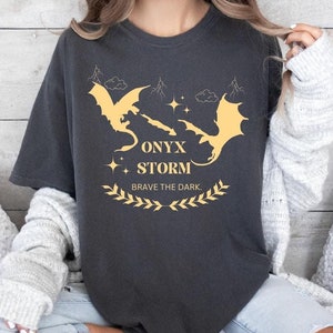 May include: A dark grey t-shirt with a graphic of two gold dragons and the text "ONYX STORM" and "BRAVE THE DARK".