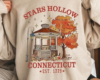 May include: A beige sweatshirt with a graphic print of a gazebo with a red roof and a fall-colored tree. The text "Stars Hollow Connecticut *EST. 1779*" is printed below the image.
