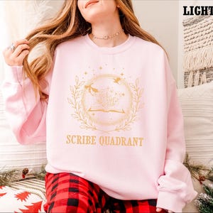 May include: Light pink sweatshirt with a gold design featuring an open book, birds, and botanical elements. The text "SCRIBE QUADRANT" is below the design. The sweatshirt is part of a clothing collection.