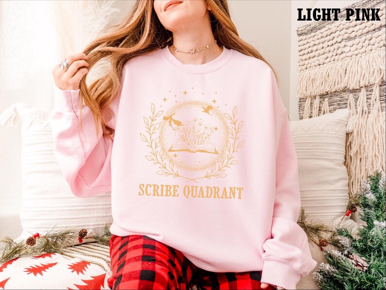 May include: Light pink sweatshirt with a gold design featuring an open book, birds, and botanical elements. The text "SCRIBE QUADRANT" is below the design. The sweatshirt is part of a clothing collection.