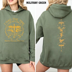 Puede incluir: Una sudadera con capucha verde con un estampado gr&aacute;fico amarillo en la parte delantera que dice "Tuve el mejor momento de mi vida. Luchando contra dragones contigo." La parte posterior de la sudadera tiene una l&iacute;nea vertical de siluetas de dragones amarillos.