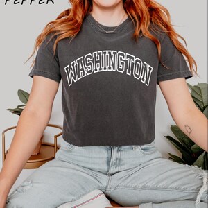 May include: A black cropped t-shirt with the word "WASHINGTON" printed in white on the front. The shirt is being worn by a person with long red hair and is paired with light blue jeans.