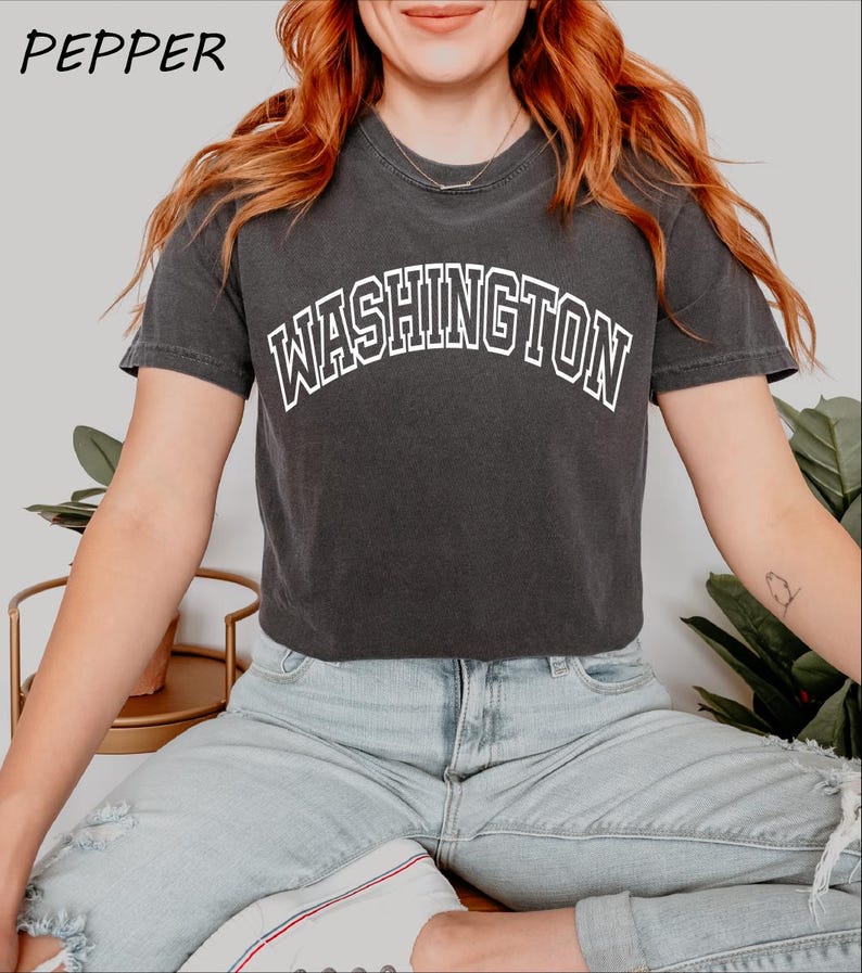 May include: A black cropped t-shirt with the word "WASHINGTON" printed in white on the front. The shirt is being worn by a person with long red hair and is paired with light blue jeans.