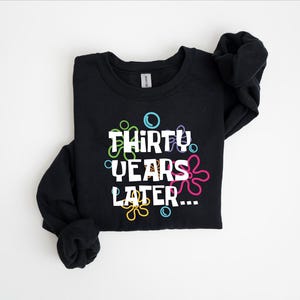 Puede incluir: Sudadera negra con un gr&aacute;fico blanco que dice "Thirty Years Later..." rodeado de burbujas de dibujos animados de colores.
