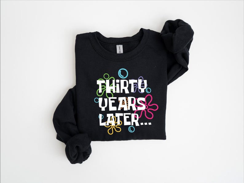 Puede incluir: Sudadera negra con un gr&aacute;fico blanco que dice "Thirty Years Later..." rodeado de burbujas de dibujos animados de colores.