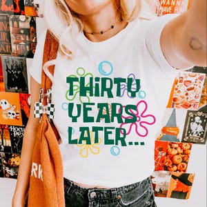 Puede incluir: Camiseta blanca con un gr&aacute;fico verde que dice "Thirty Years Later..." rodeado de burbujas y flores de dibujos animados de colores.