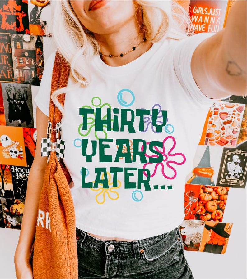 Puede incluir: Camiseta blanca con un gr&aacute;fico verde que dice "Thirty Years Later..." rodeado de burbujas y flores de dibujos animados de colores.