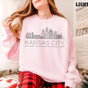 May include: A light pink sweatshirt with a black outline of the Kansas City skyline and the text "KANSAS CITY".
