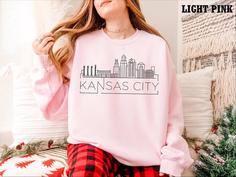 May include: A light pink sweatshirt with a black outline of the Kansas City skyline and the text "KANSAS CITY".