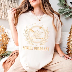May include: Ivory-colored t-shirt with a gold-colored circular graphic featuring an open book, leaves, and birds. The text "SCRIBE QUADRANT" is below the graphic. The shirt is part of a clothing collection.