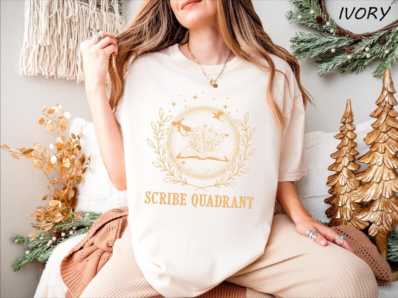 May include: Ivory-colored t-shirt with a gold-colored circular graphic featuring an open book, leaves, and birds. The text "SCRIBE QUADRANT" is below the graphic. The shirt is part of a clothing collection.