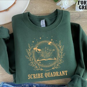 May include: Forest green sweatshirt with a circular design featuring an open book, flowers, and flying figures. The text "SCRIBE QUADRANT" is printed below the design. The sweatshirt is folded, with the sleeves rolled up.