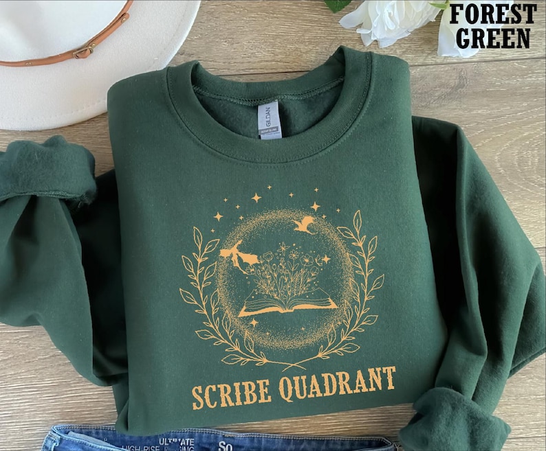 May include: Forest green sweatshirt with a circular design featuring an open book, flowers, and flying figures. The text "SCRIBE QUADRANT" is printed below the design. The sweatshirt is folded, with the sleeves rolled up.