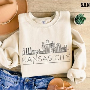 May include: A beige sweatshirt with a black outline of the Kansas City skyline and the text "KANSAS CITY" printed on the front. The sweatshirt is laid out on a wooden surface with a pair of jeans, a hat, and a potted plant.