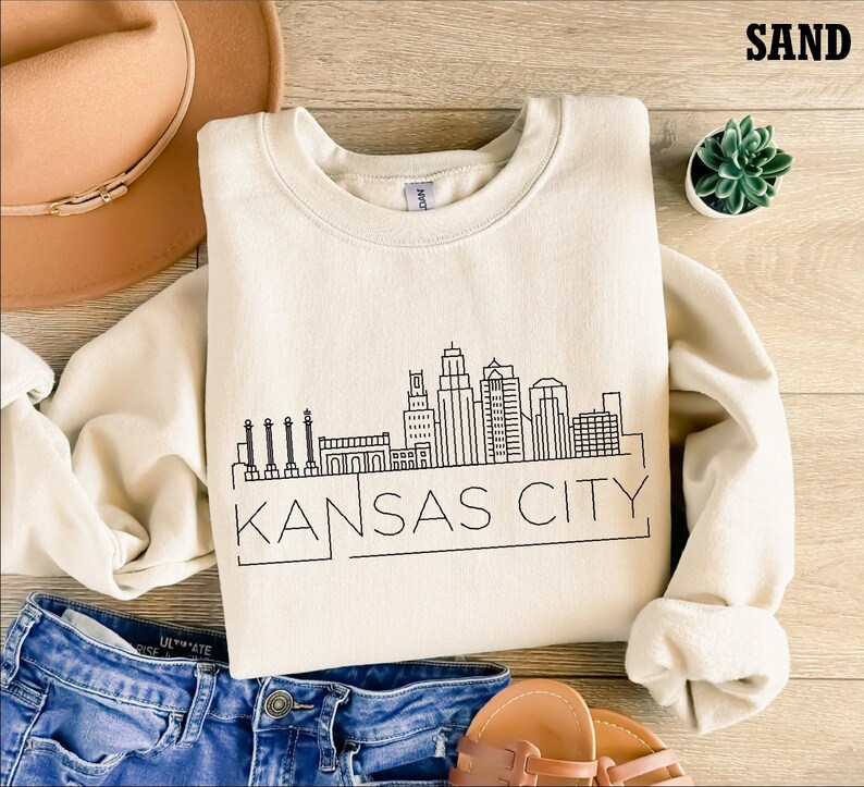 May include: A beige sweatshirt with a black outline of the Kansas City skyline and the text "KANSAS CITY" printed on the front. The sweatshirt is laid out on a wooden surface with a pair of jeans, a hat, and a potted plant.