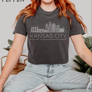 May include: A dark gray cropped t-shirt with a white outline of the Kansas City skyline and the text "KANSAS CITY".