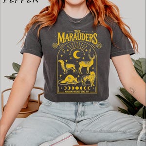 May include: A black t-shirt with a yellow graphic design featuring the text "The Marauders" and a crescent moon, stars, and animals. The text below the graphic reads "Managing Mischief Since 1971."