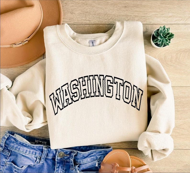 May include: A cream-colored sweatshirt with the word "WASHINGTON" printed in black across the front. The sweatshirt is laid out on a wooden surface with a brown hat and a small potted plant.