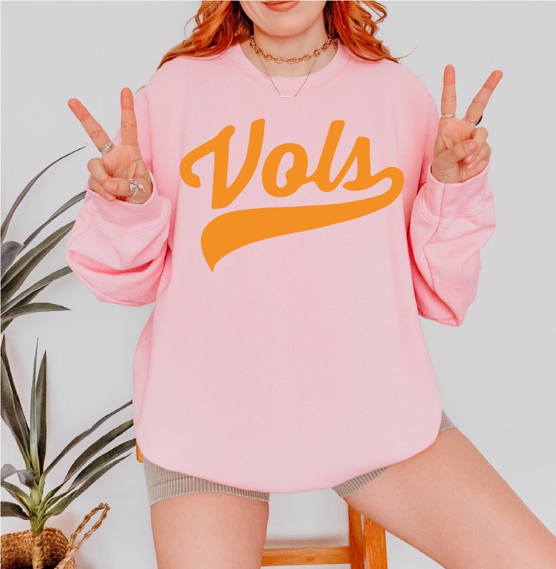 May include: A light pink crewneck sweatshirt featuring the word Vols printed in a bold orange script font across the chest. The garment has long sleeves with ribbed cuffs and a ribbed neckline, providing a simple and clean design for casual wear.