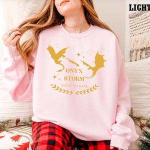 May include: A light pink sweatshirt featuring a graphic of two golden dragons and the text "ONYX STORM BRAVE THE DARK".