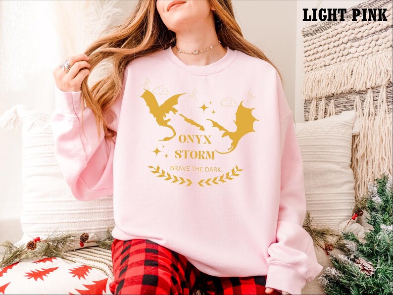 May include: A light pink sweatshirt featuring a graphic of two golden dragons and the text "ONYX STORM BRAVE THE DARK".