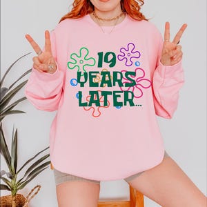 May include: Pink sweatshirt with a green graphic that says "19 years later..." surrounded by colorful cartoon flowers.