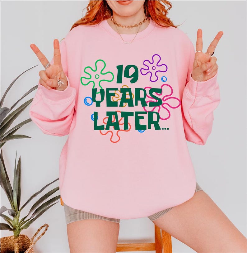 May include: Pink sweatshirt with a green graphic that says "19 years later..." surrounded by colorful cartoon flowers.