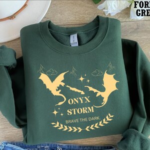 May include: Forest green sweatshirt featuring a graphic of two yellow dragons and the text "Onyx Storm Brave the Dark."
