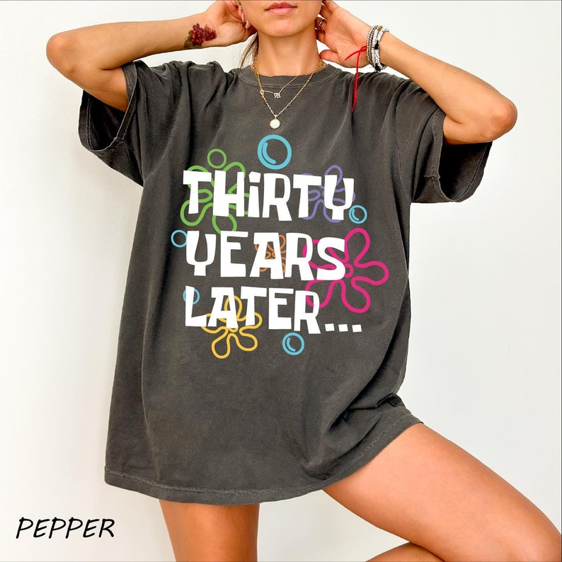 Puede incluir: Una camiseta de gran tama&ntilde;o de color gris oscuro con el texto "Thirty Years Later..." en blanco, rodeada de flores y burbujas de dibujos animados de colores.