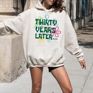 Puede incluir: Una sudadera con capucha blanca con un gr&aacute;fico verde que dice "Thirty Years Later..." con flores de dibujos animados de colores alrededor del texto.