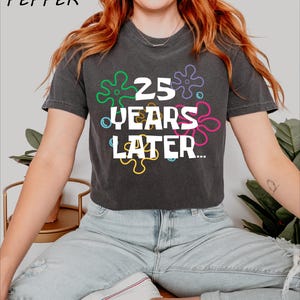 May include: A black cropped t-shirt with a white graphic that says "25 Years Later..." surrounded by colorful cartoon-style flowers.