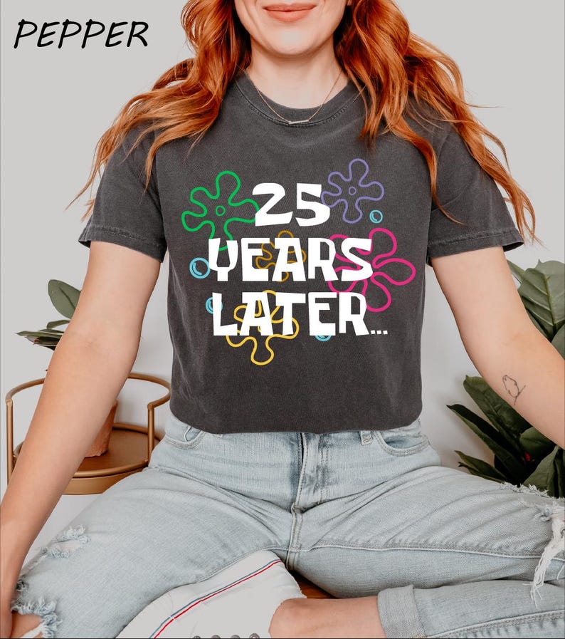May include: A black cropped t-shirt with a white graphic that says "25 Years Later..." surrounded by colorful cartoon-style flowers.