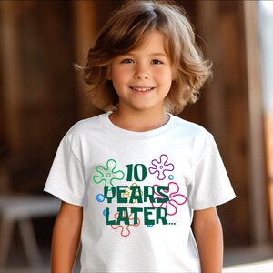 May include: White t-shirt with a green graphic that says "10 Years Later" surrounded by colorful cartoon flowers.
