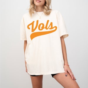May include: An off-white, short-sleeved t-shirt featuring the word "Vols" printed in a bold, orange, cursive script across the chest. The shirt has a crew neckline and a relaxed, loose-fitting silhouette that extends to the upper thigh area.