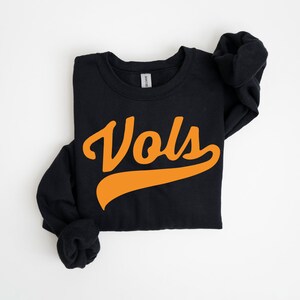 May include: A black long-sleeve crewneck sweatshirt featuring the word Vols printed in a bold orange script font across the center chest. The garment has a ribbed neckline and cuffs, with a standard fit and a visible brand tag at the inner collar.