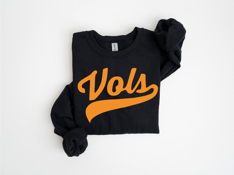 May include: A black long-sleeve crewneck sweatshirt featuring the word Vols printed in a bold orange script font across the center chest. The garment has a ribbed neckline and cuffs, with a standard fit and a visible brand tag at the inner collar.