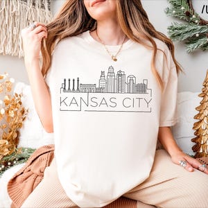 May include: A white t-shirt with a black outline of the Kansas City skyline and the text "KANSAS CITY". The shirt is being modeled by a person wearing beige pants.