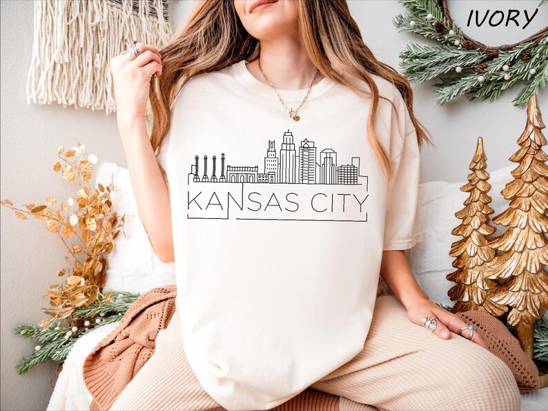 May include: A white t-shirt with a black outline of the Kansas City skyline and the text "KANSAS CITY". The shirt is being modeled by a person wearing beige pants.