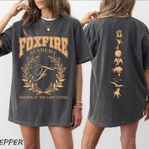 May include: Dark gray t-shirt with "FOXFIRE ACADEMY" in orange block letters, a laurel wreath, and the text "KEEPER OF THE LOST CITIES." The back features orange illustrations of mythical creatures. Apparel.