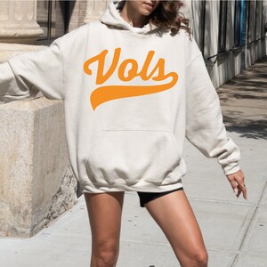 May include: An oversized cream-colored hooded sweatshirt featuring a large orange graphic on the chest with the text "Vols" in a cursive script font. The garment includes long sleeves, a front kangaroo pocket, and a drawstring hood.